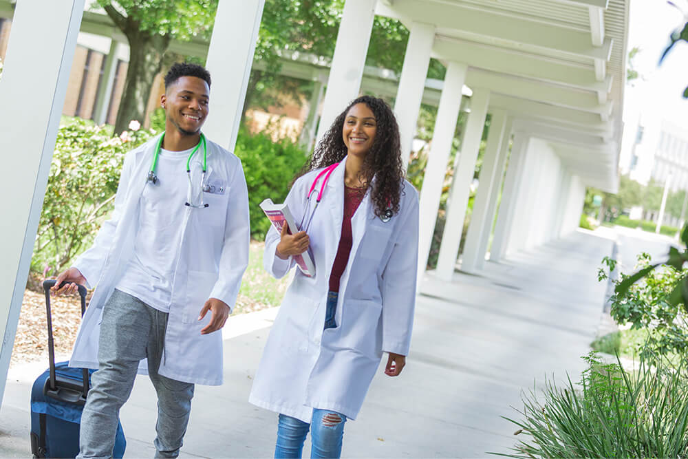 Two nursing students wearing stethescopes walking down an exterior corridor